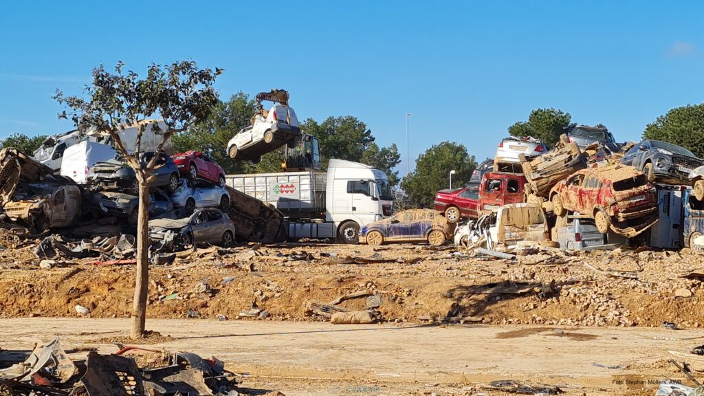 Schlammige Fläche mit Baum vorne, beschmutzte und beschädigte Fahrzeuge auf einem Haufen rechts, links Verladung eine Fahrzeugs in einen Container LKW