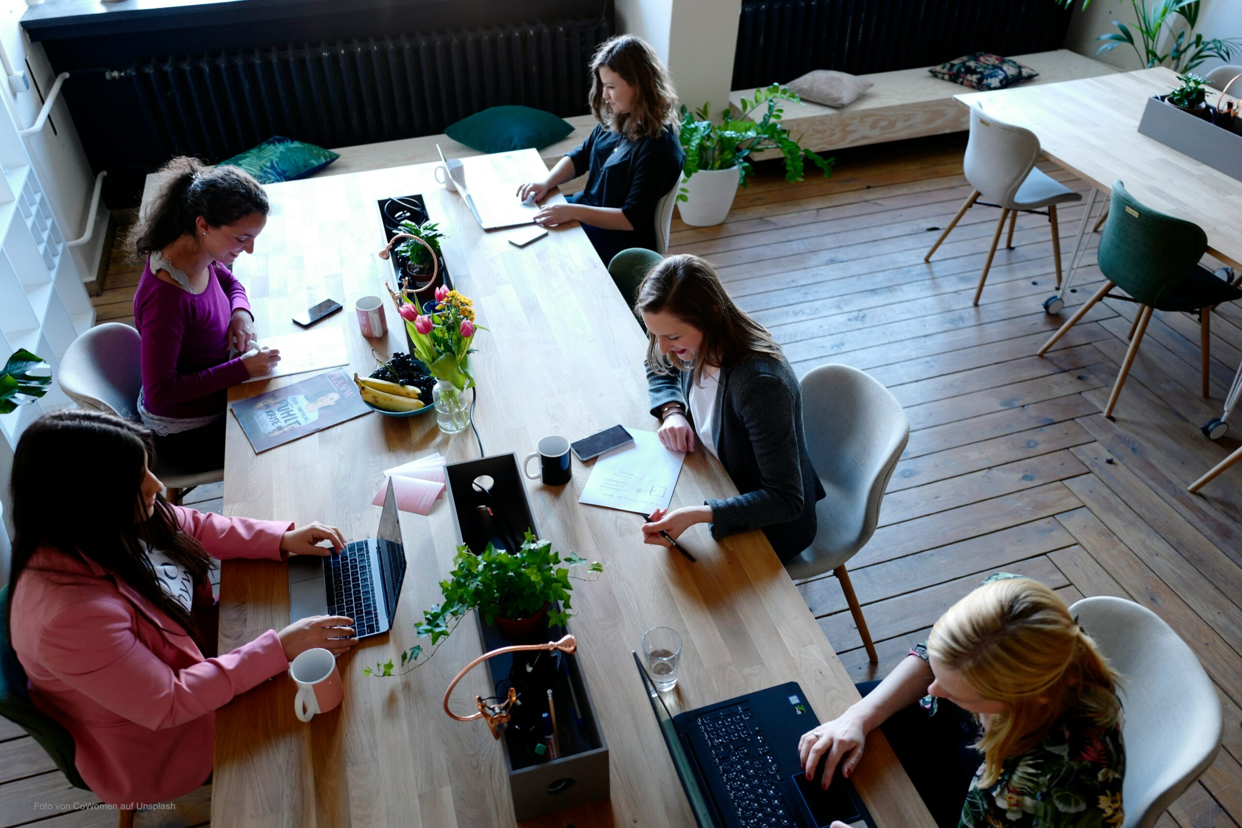 Arbeitstisch links mit vier Personen sitzend mit Computer und Unterlagen. Blumen in der Mitte.